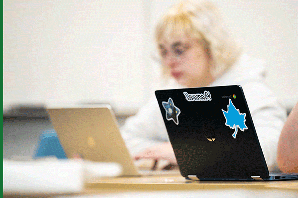 A black laptop is visible in the foreground on a table in a classroom. It has a blue Sycamore leaf sticker with a white outline on the top left, and another stick top center that says Cromwell. A third sticker, further right, is shaped like a star. Out of focus in the background, a blonde female student wearing glasses and a white hoodie can be seen typing on a silver laptop.  